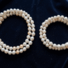 A richly lit flat-lay of lustrous white and cream Bahraini natural pearls arranged on dark navy velvet fabric. A delicate pearl necklace and matching pearl bracelet are placed side by side. Soft golden warm light from the upper left creates gentle highlights on each pearl's surface, revealing a soft pink and silver overtone. The composition is elegant and minimal, with a shallow depth of field blurring the background slightly. No text or words appear anywhere in the image.