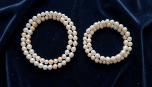 A richly lit flat-lay of lustrous white and cream Bahraini natural pearls arranged on dark navy velvet fabric. A delicate pearl necklace and matching pearl bracelet are placed side by side. Soft golden warm light from the upper left creates gentle highlights on each pearl's surface, revealing a soft pink and silver overtone. The composition is elegant and minimal, with a shallow depth of field blurring the background slightly. No text or words appear anywhere in the image.